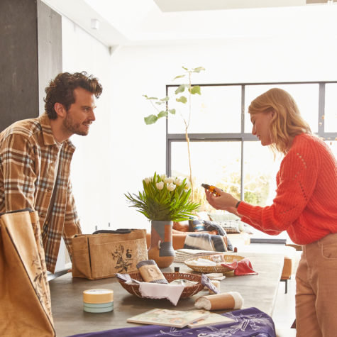 Un homme et une femme discutant dans une pièce lumineuse, la femme montrant un objet à l'homme sur une table avec divers articles H2O at Home et une plante en vase